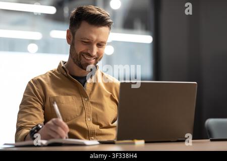 Smiling Male Professional Writing In Notebook, Working On Laptop At Modern Office Stock Photo