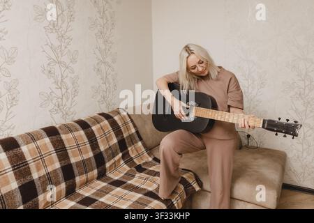 A mature woman sits comfortably on a couch and practices playing an acoustic guitar at home. Stock Photo