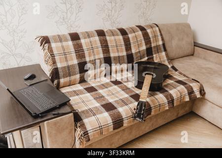 A black acoustic guitar rests on a couch beside an open laptop, smartphone and computer mouse in a living room. Stock Photo