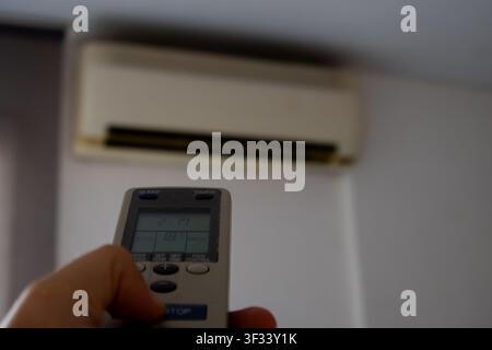 Close-up of a person's hand pointing a remote control toward a wall-mounted air conditioner inside a home. The image represents indoor climate control Stock Photo