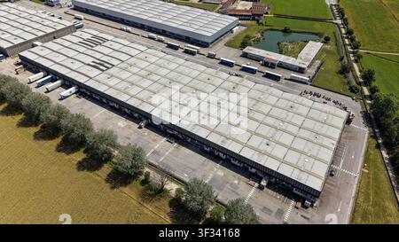 Aerial view of a large industrial logistics warehouse complex featuring loading docks, parked semi-trucks, and surrounding green fields in Caleppio, L Stock Photo