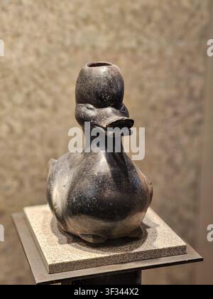 Metallic duck sculpture sits on a stone pedestal. Smooth surface reflects subtle light and shadow. Dark gray finish gives it an ancient, weathered loo Stock Photo