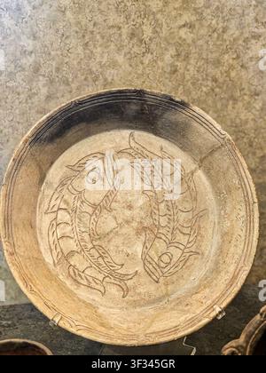 Ancient clay bowl rests on stone surface. Etched fish swirl in circular pattern. Brownish tones show age and use. Rough edges hint at handcrafted orig Stock Photo