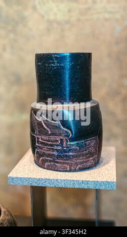 Dark ceramic vessel stands on display. Smooth surface reflects subtle light. Carved details suggest ancient craftsmanship. Neutral tones evoke quiet m Stock Photo