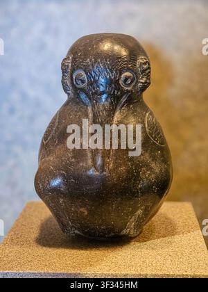 Ancient carved stone figure sits solemnly on display. Dark surface reflects subtle light, emphasizing its form. Smooth curves and stylized features su Stock Photo
