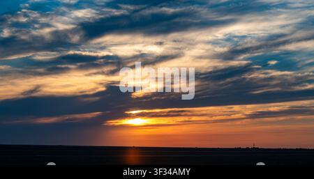 A dramatic sunset over a flat horizon. Vibrant orange and golden sunbeams break through layered clouds in a darkening blue sky. A scenic natural backg Stock Photo