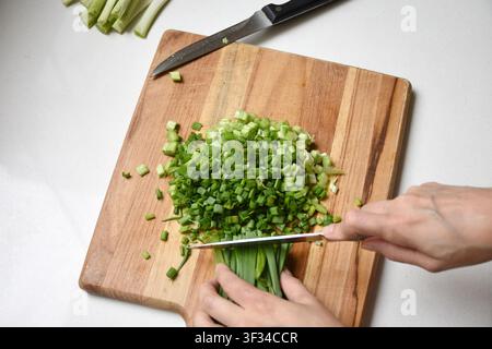 A close-up of a hand slicing fresh green onions on a rustic wooden cutting board. Natural lighting highlights the vibrant color and texture, creating Stock Photo