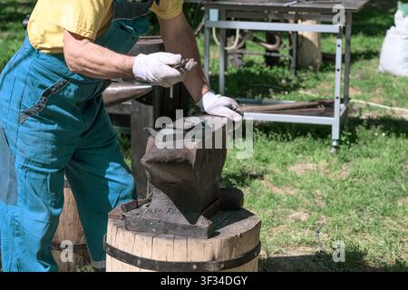 A blacksmith wearing protective gloves handles a small metal piece on a heavy anvil at an outdoor metalworking setup. Stock Photo