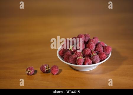 A close-up horizontal photograph of a small white ceramic bowl filled to the brim with fresh red raspberries. Three individual berries lie on the smoo Stock Photo