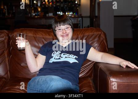 A smiling young woman with Down syndrome holding a glass of water while relaxing on a leather sofa in a pub interior Stock Photo