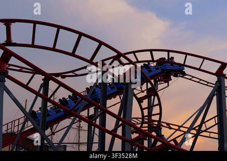 Roller coaster cars filled with people moving quickly along a red track against a sky with soft orange and pink clouds during sunset, creating an exci Stock Photo