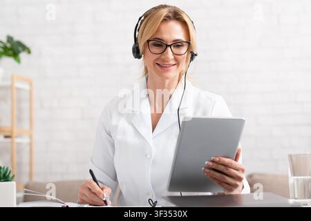 Visit to doctor remotely. Smiling doctor in glasses and headphones holds digital tablet in hands Stock Photo