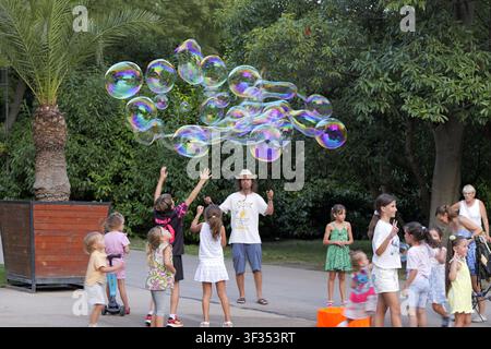 Children of various ages are reaching for large soap bubbles created by a street performer during a sunny day in a park. Bulgaria, varna, 20 july 2026 Stock Photo