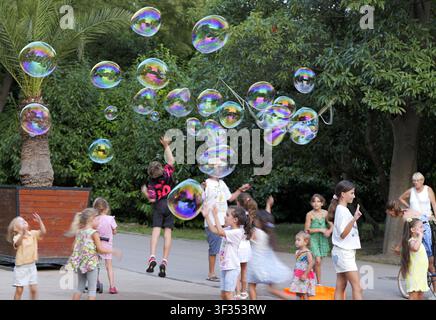 Children running and reaching for large colorful soap bubbles floating in a green park during a sunny summer day. Bulgaria, varna, 20 july 2026 Stock Photo