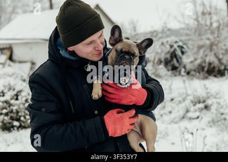 A man stands in a snowy yard while holding a small dog. The man wears gloves and a winter jacket. The dog is wearing a coat. Snow covers the ground an Stock Photo