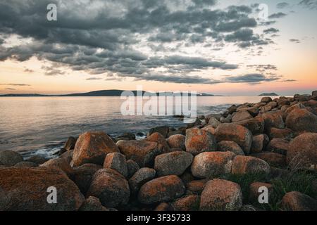 View of the rocky coastline at Middleton Beach with orange-lichen-covered boulders, calm ocean waters, and a dramatic cloudy sky at sunset in Middleto Stock Photo