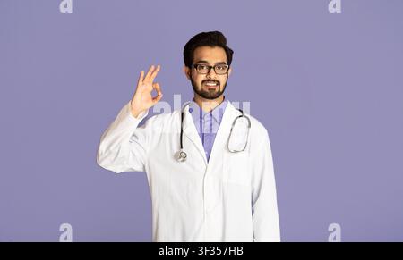 Positive Indian doctor showing OK gesture on lilac background Stock Photo