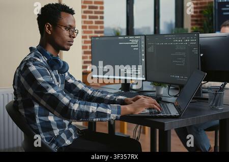 Programmer thinking while touching beard and fixing glasses while typing on laptop sitting at desk with multiple screens parsing code around computer screens, showing developer teamwork, source. Stock Photo