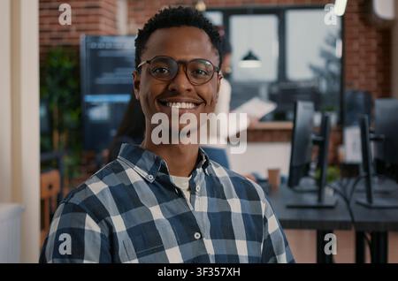 Focused portrait with african american programmer sitting down working on laptop arranging glasses looking up and smiling at camera around computer screens, showing developer teamwork, source code. Stock Photo