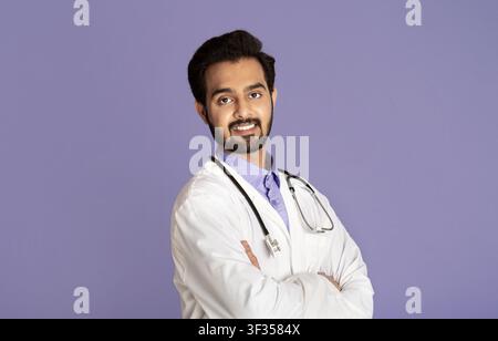 Smiling Indian doctor in lab coat standing with crossed arms on violet background Stock Photo