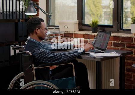 Wheelchair worker plans business growth online at a workplace computer while creating software, writing algorithms and supporting cloud computing development in an IT office. Stock Photo