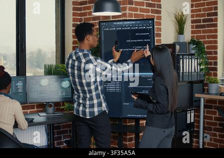 Software engineers analyzing code on wall screen tv comparing errors using digital tablet next to programmer working with artificial intelligence, with artificial intelligence and big data. Stock Photo