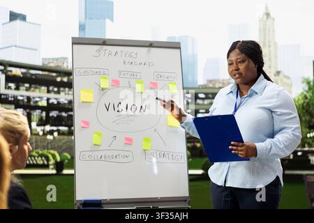 African american woman presenter leading outdoor meeting with vision board, explaining roadmap presentation and sharing ideas for project overview. Speaking for business team. Stock Photo