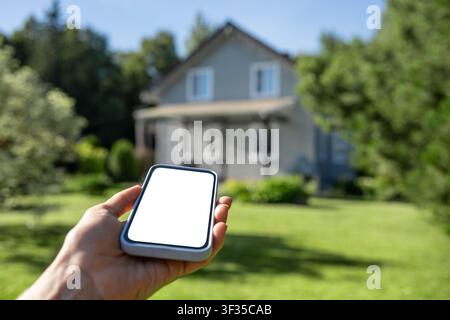 Hand holding smartphone with white screen against modern house blurred in background. Concept of smart home, real estate and digital property manageme Stock Photo