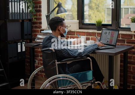 Wheelchair worker plans a financial business project on a laptop while wearing a face mask, linking accessible office work, chronic disability and startup planning. Stock Photo