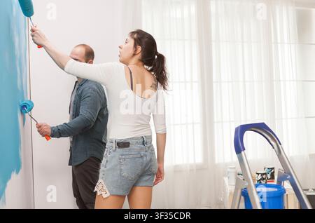 Young family painting living room wall together in living room, showing painting tools, home repair and apartment improvement, showing hands-on creative work, tools and practical progress. Stock Photo