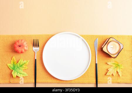 Autumn table setting with white plate, golden cutlery, candle, maple leaves and decorative pumpkin on beige background. Top view, flat lay, copy space Stock Photo