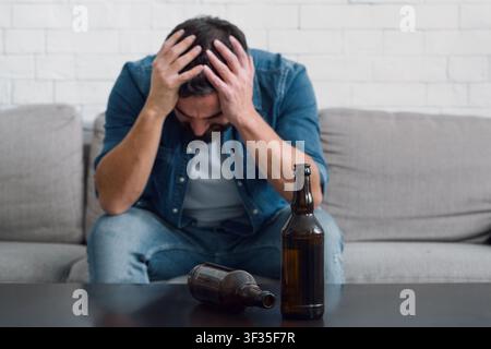 Sad man holds his head and sitting on couch, empty bottles on table, close up Stock Photo