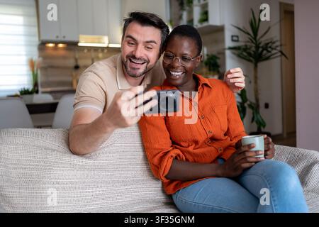 A man and a woman sit on a couch in their living room and look at a smartphone. Stock Photo