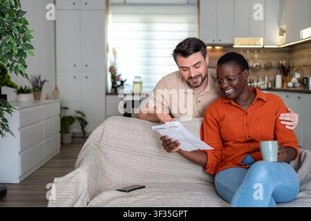 A man and a woman sit together on a sofa in their home. They look at documents and share a relaxed moment. The woman holds a cup. Stock Photo