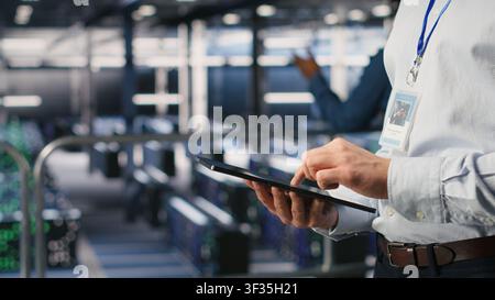 Close up of server room worker checking code on tablet next to colleague reviewing AI LLM visualization. Data center IT expert using device to monitor artificial intelligence systems Stock Photo