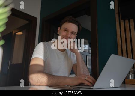 Thoughtful young man rests his chin on his hand beside a laptop while working remotely in a modern apartment in Prague, Czech Republic. Stock Photo