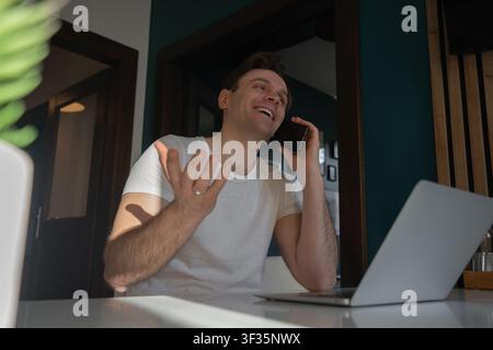 Happy young man talks on a smartphone and gestures beside a laptop while working remotely in an apartment in Prague, Czech Republic. Stock Photo