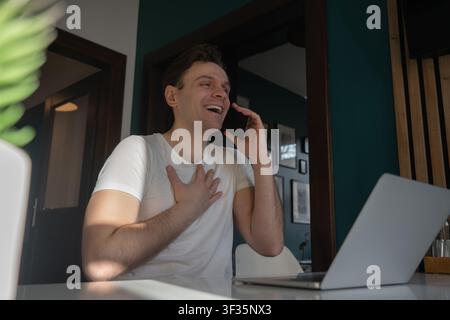 Happy young man places a hand on his chest while reacting with gratitude during a smartphone call beside a laptop in Prague, Czech Republic. Stock Photo