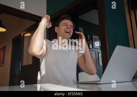 Excited young man raises his fist in celebration while talking on a smartphone beside a laptop in Prague, Czech Republic. Stock Photo