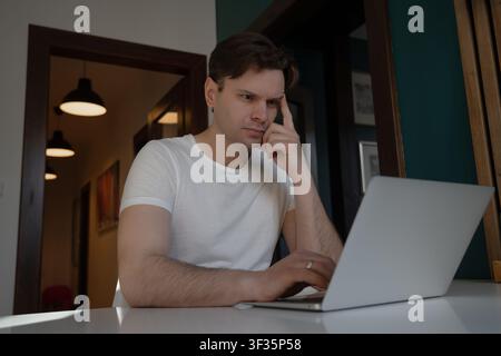 Thoughtful young man touches his temple while solving a difficult problem on a laptop in Prague, Czech Republic. Stock Photo