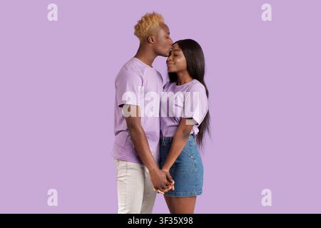 Tender black man giving his girlfriend kiss on forehead, expressing his affection on violet studio background Stock Photo