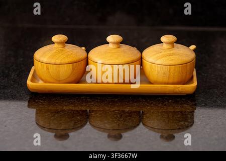 Three round bamboo jars with lids on a matching tray. Set on a dark reflective surface. Minimalist wooden kitchenware design for spices or condiments Stock Photo
