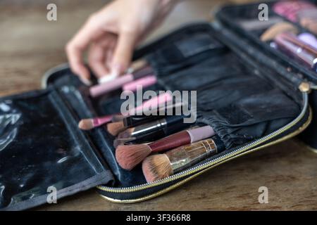 A makeup brush set in black zippered case on wooden table, hand reaching for brushes, soft natural light and organized cosmetic tools conveying care a Stock Photo
