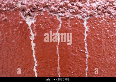 A red painted concrete wall with vertical white mineral streaks and rough textured surface showing weathered damage and peeling layer Stock Photo