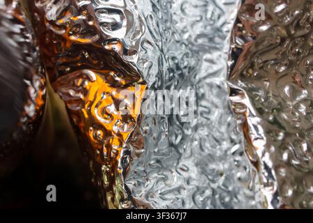 A shiny textured metallic foil with amber reflection and waterlike ripples creating abstract reflective pattern and soft highlights Stock Photo