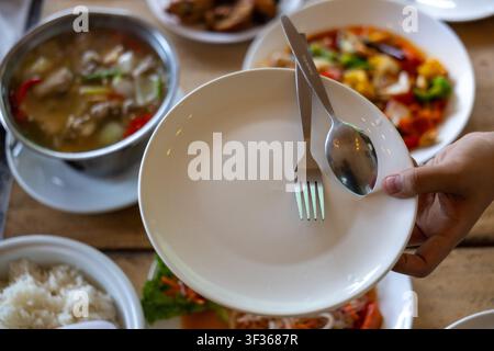 A white empty ceramic plate held by hands with fork and spoon ready over table set with asian dishes rice soup and stir fry, inviting communal dining Stock Photo