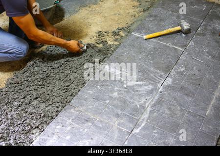 A worker spreading cement mortar with trowel while laying floor tile, focused hands and tools creating textured surface and practical construction sce Stock Photo