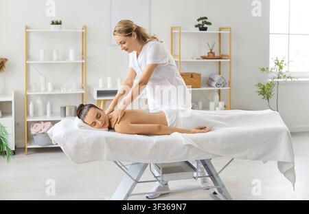 Female masseur giving relaxing back and shoulder massage to young woman client in spa salon. Stock Photo