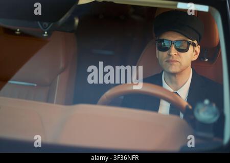 Caucasian middle aged chauffeur driving luxury car while wearing sunglasses and cap, conveying premium transport service. Ideal for executive travel, private transfer marketing, close up Stock Photo