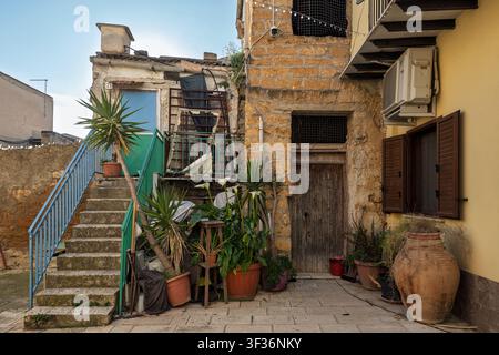 A rustic courtyard of a stone house featuring outdoor stairs, various potted plants, and an aged wooden door, Agrigento, Sicily Stock Photo